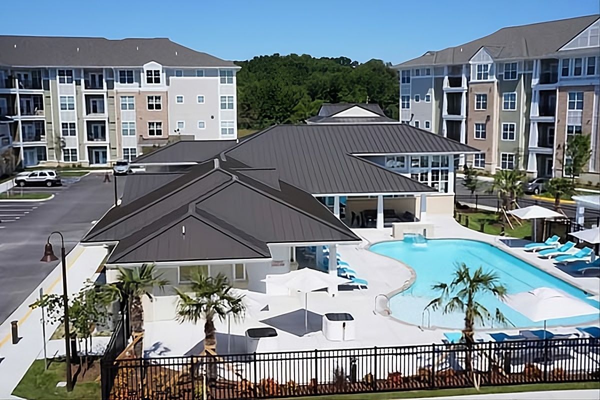 An aerial view of an apartment complex featuring a swimming pool, lounge area with palm trees, and multi-story buildings.