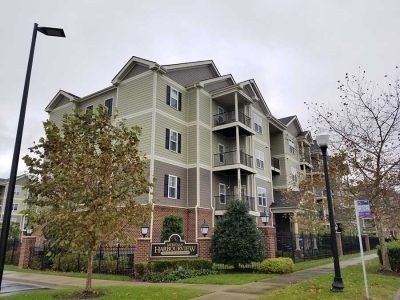 Apartment building with green and brown siding, balconies, and a sign in front. Overcast sky.