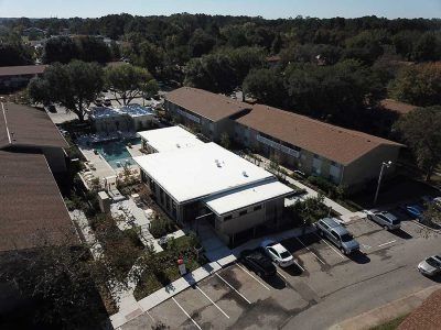 Aerial view of an apartment complex with a pool, parking lot, and surrounding trees. Brown roofs, white building.