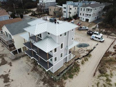 Gray beach house with balconies, metal roof. Vehicles parked nearby on sandy ground.