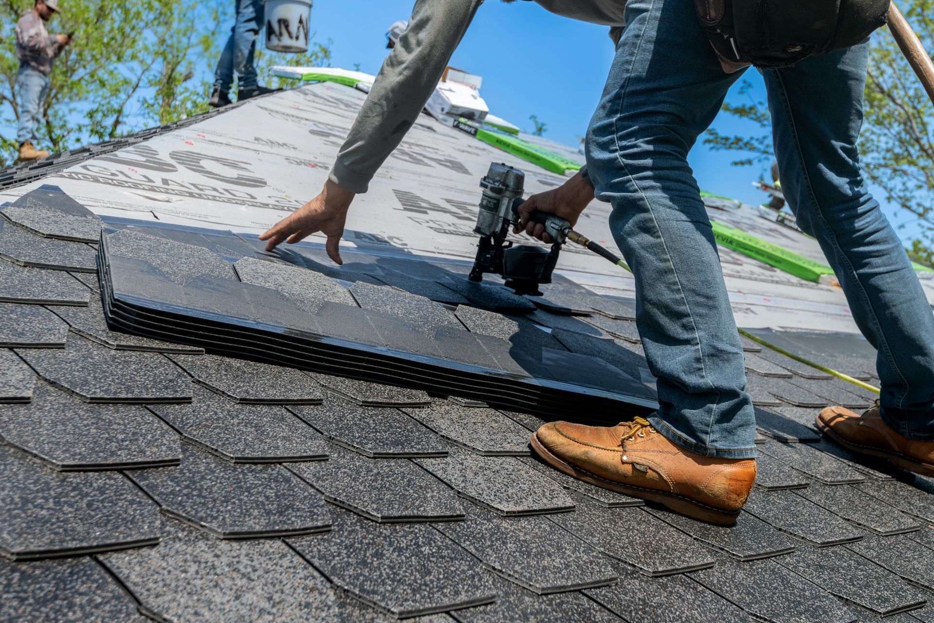 Roofer using a nail gun to install asphalt shingles on a roof under a bright blue sky.