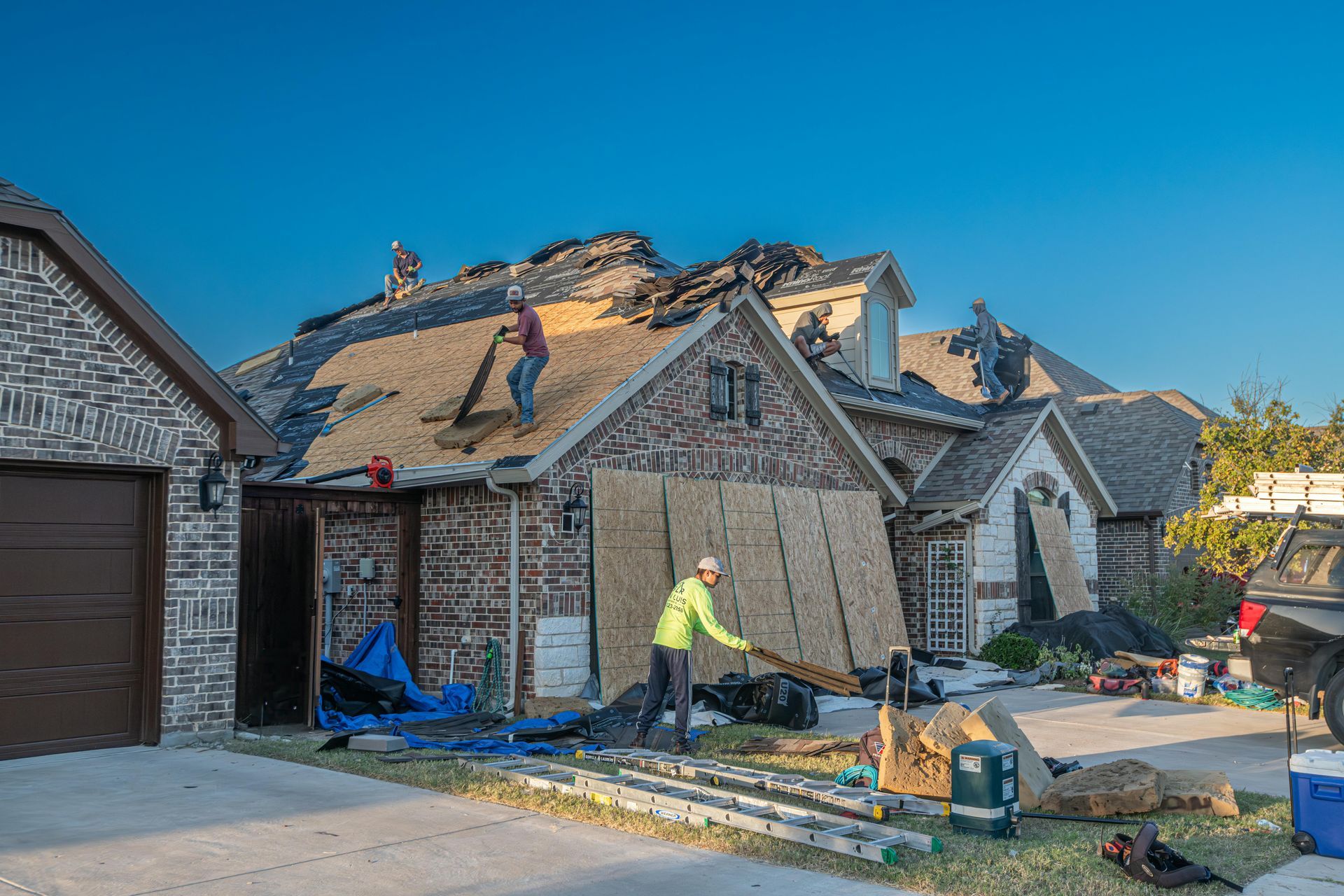 Roofers working on a house with damaged roof and exposed sheathing, blue tarp, debris on ground.
