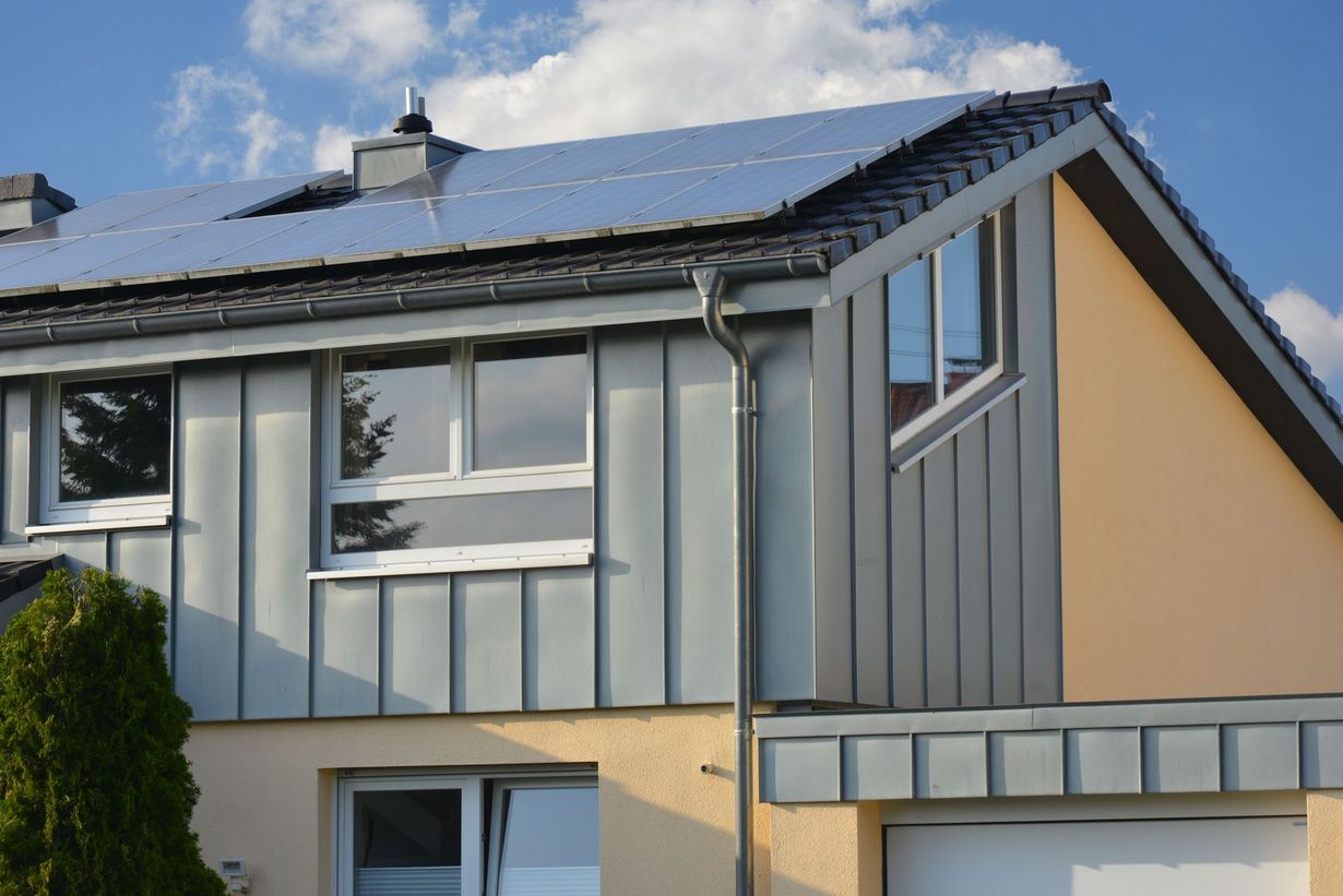 A house exterior featuring a dormer with grey metal siding, solar panels on the roof, and a beige stucco wall.
