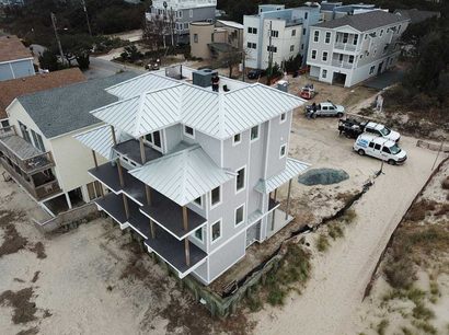 Aerial view of a multi-story light gray house with a metal roof under construction near a sandy beach and other buildings.