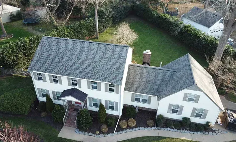 Aerial view of a white two-story suburban house with a grey shingled roof, surrounded by a lawn and mature trees.