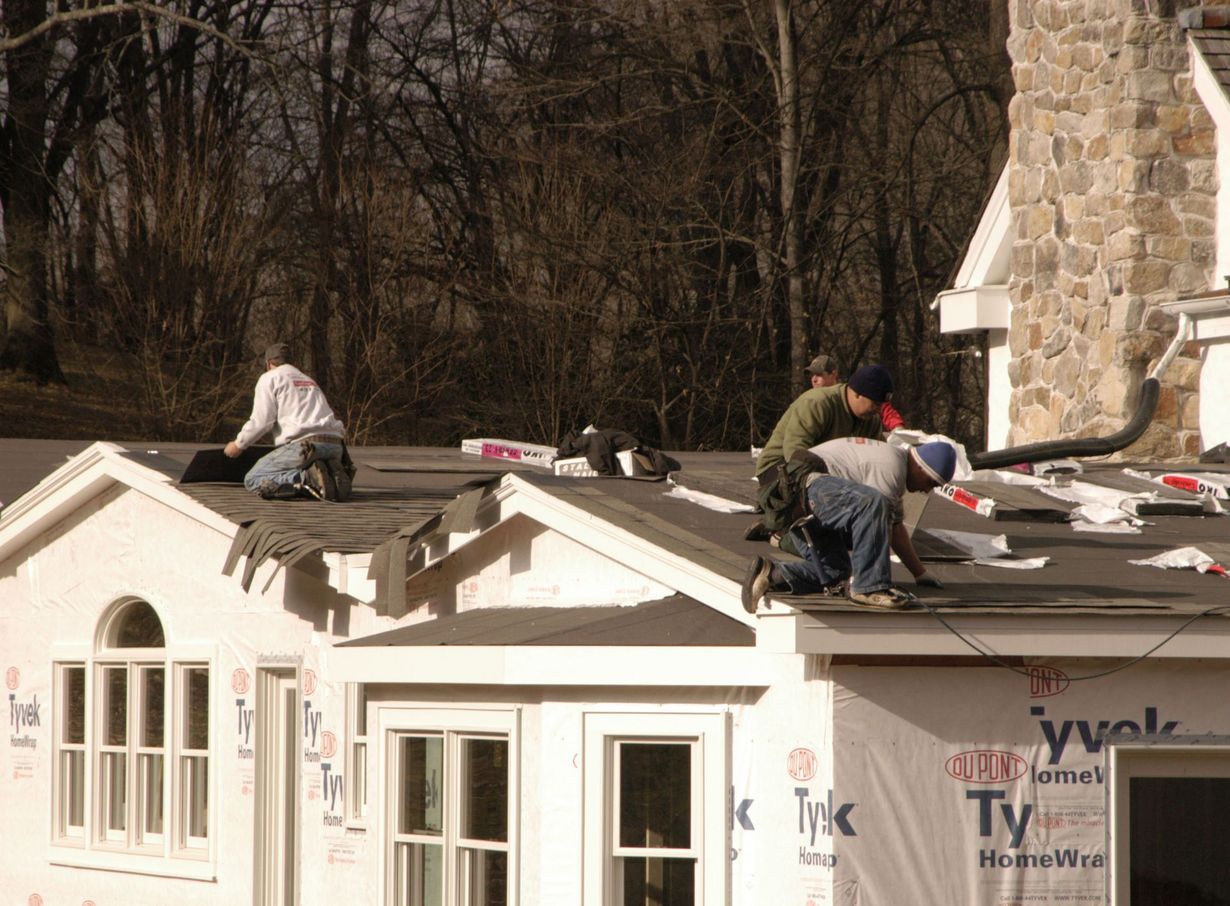 Two workers install dark underlayment on the roof of a house wrapped in white Tyvek construction paper.