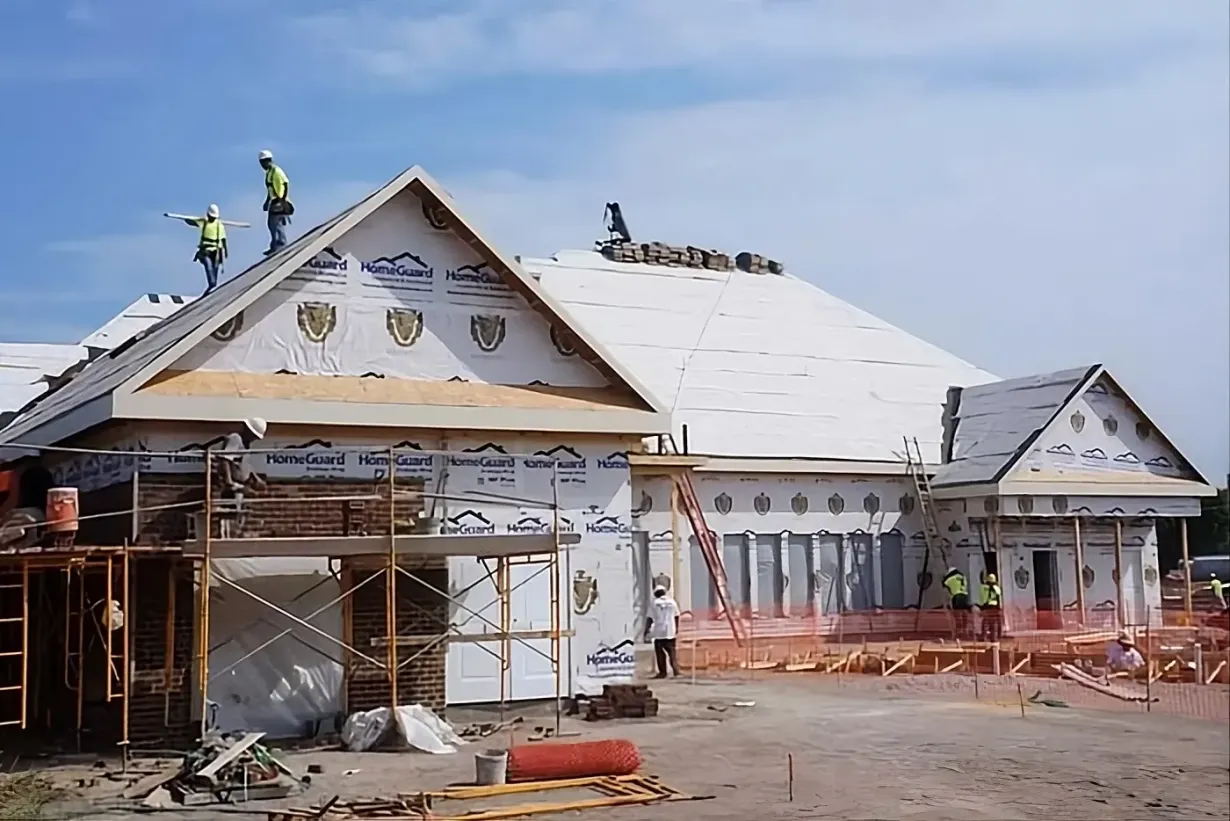 Construction workers in safety gear work on the roof and structure of a building covered in white weather-resistant wrap.
