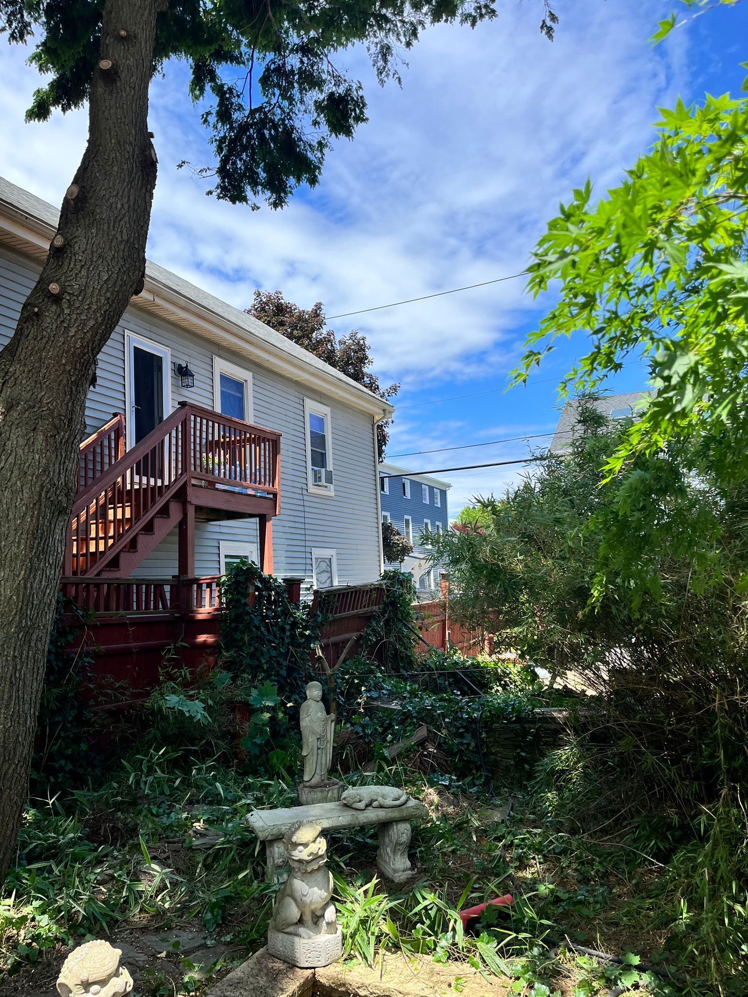 Backyard with a two-story house, wooden deck, and stone bench under a tree. Overcast day with blue sky.