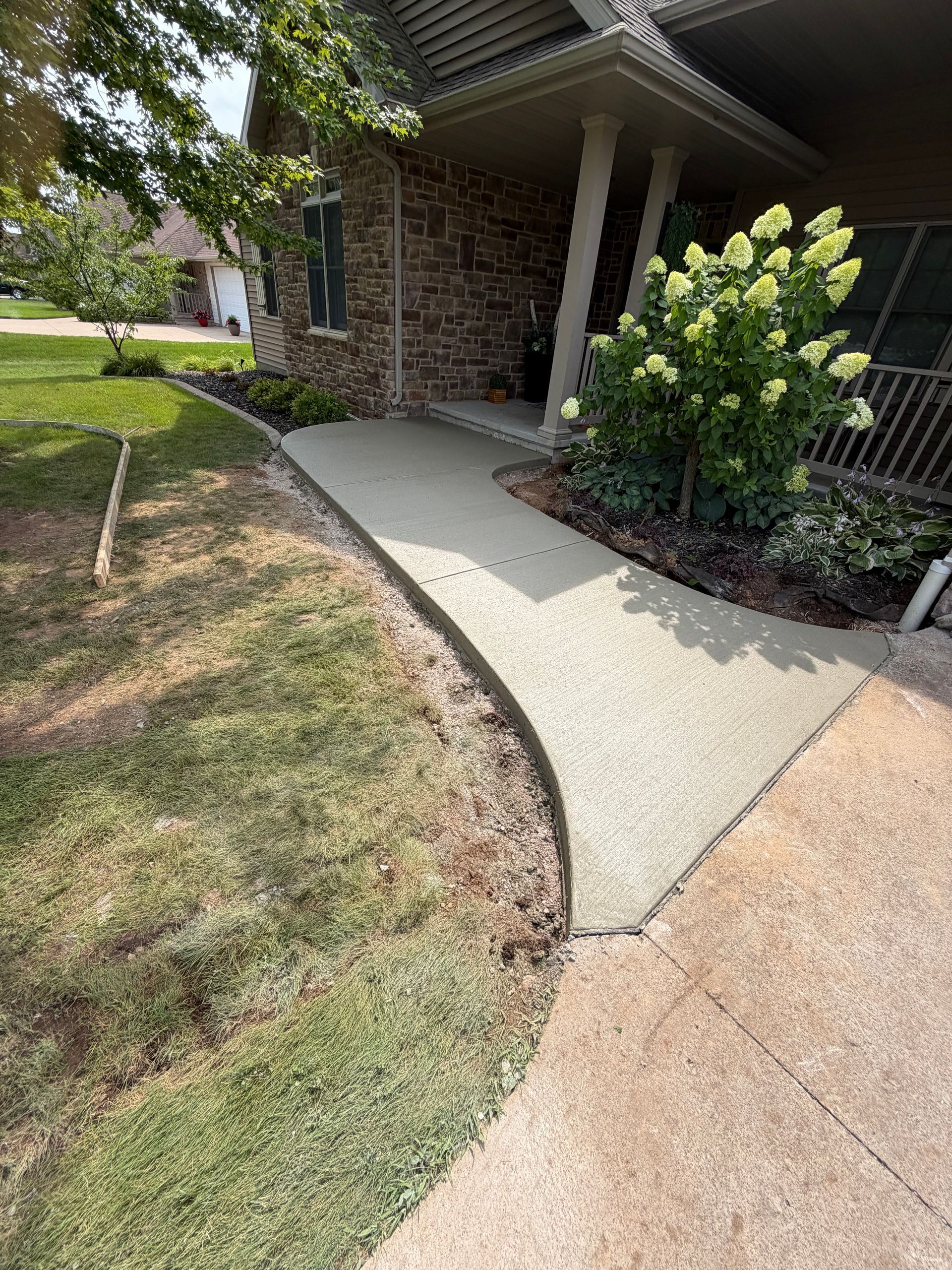 Curved concrete walkway beside a brick house and porch, bordered by grass and flowering shrubs