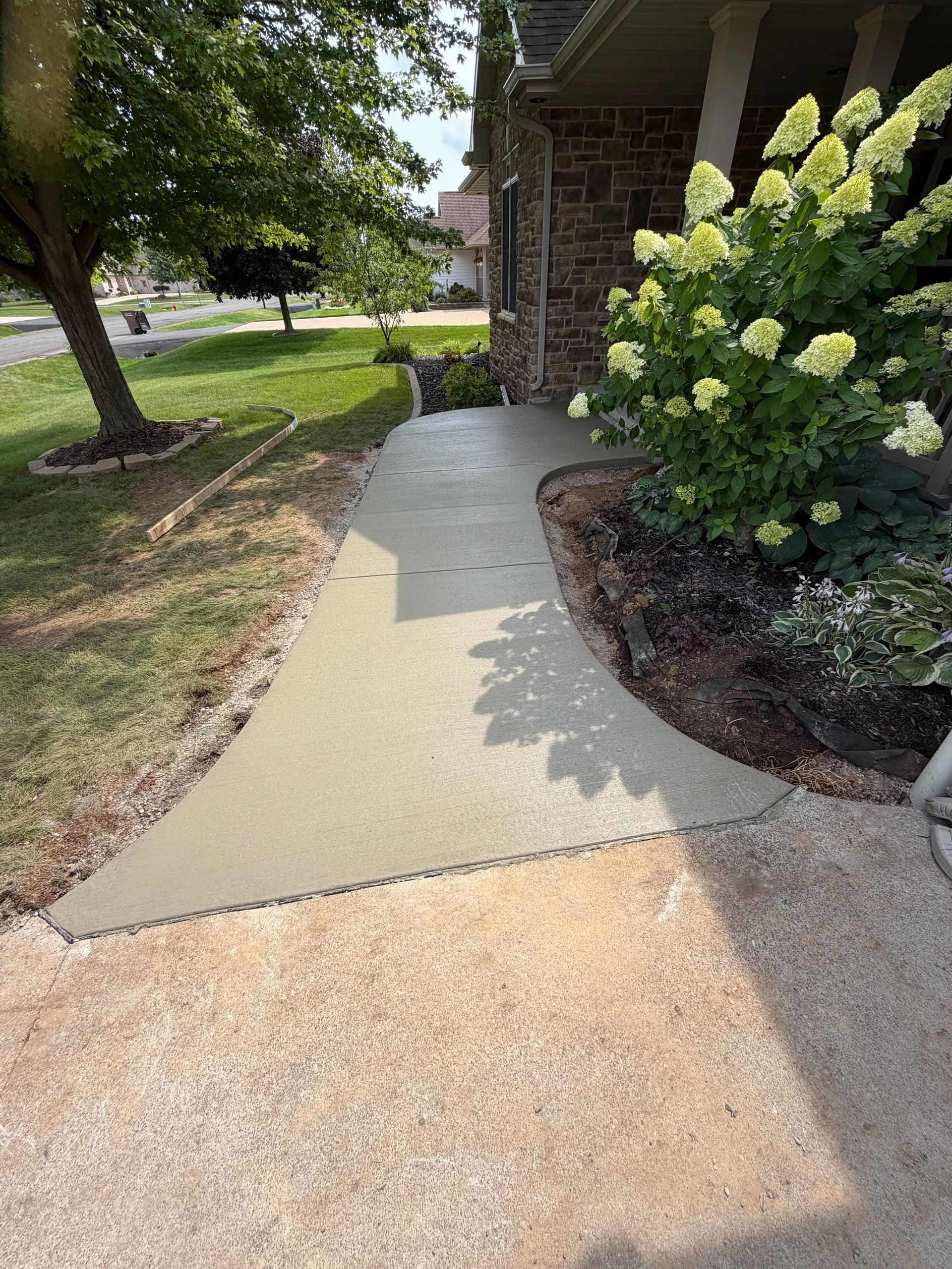 Curved concrete walkway beside a brick house, with a hydrangea bush and shaded lawn.