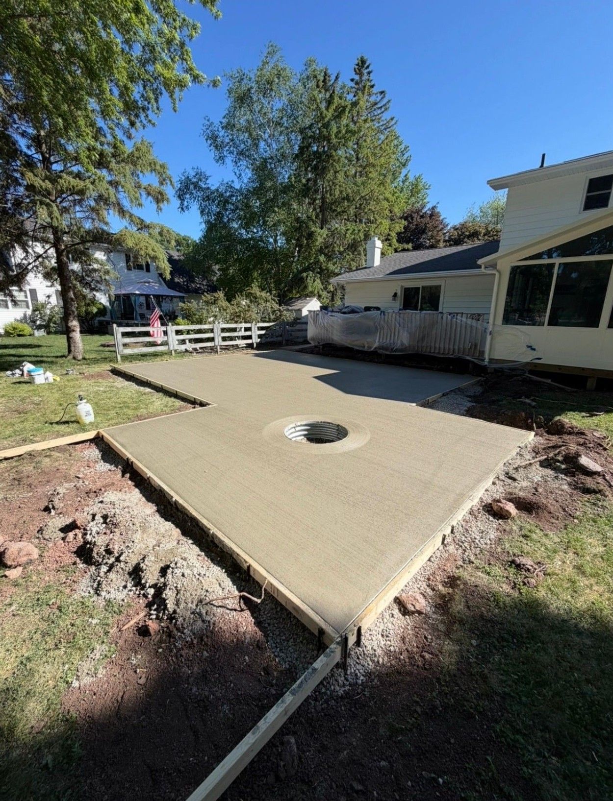 Concrete patio slab with a round drain beside a house and white fence in a sunny yard