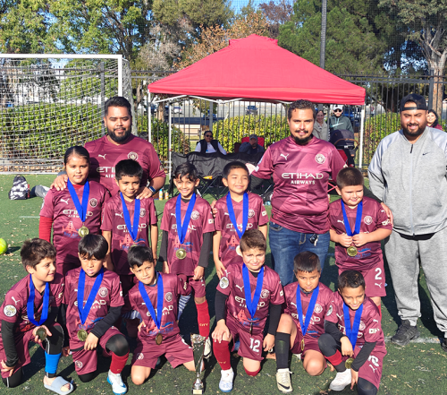 Youth soccer team in maroon uniforms pose with coaches, medals, and a trophy on a green field.
