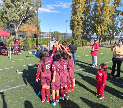 Youth soccer team huddles in maroon uniforms on a field under a sunny sky.