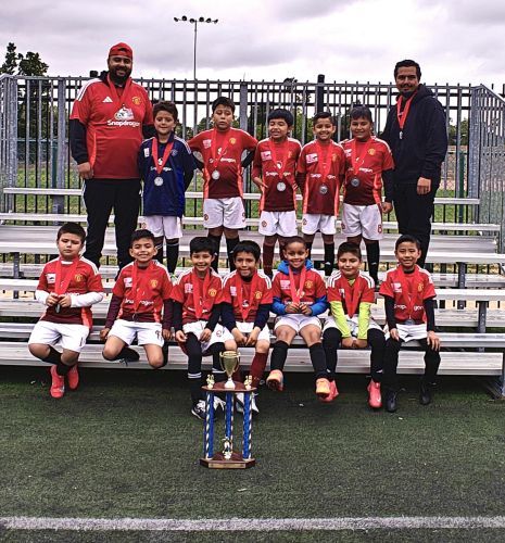 Youth soccer team in red jerseys posing with coaches and trophy on a field.