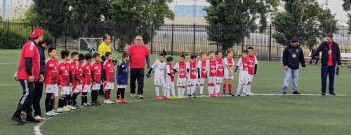 Two youth soccer teams, one in red, the other in white, line up on a green field with coaches and officials nearby.