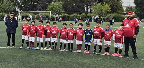 A youth soccer team wearing red jerseys poses on a green field with two coaches. Spectators in the background.