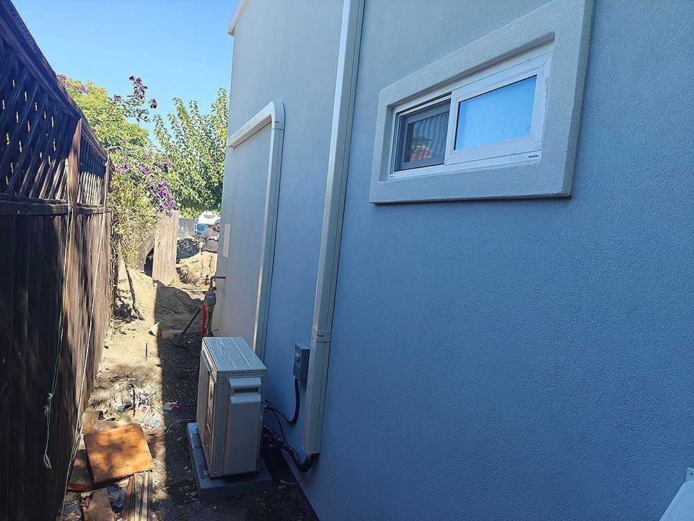 A narrow side yard with an air conditioning unit against a light blue stucco wall, next to a brown wooden fence.