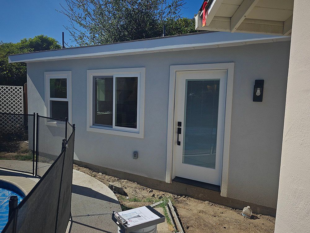 Light blue-gray building with white trim, windows, and door. Black fence and pool visible on the left.