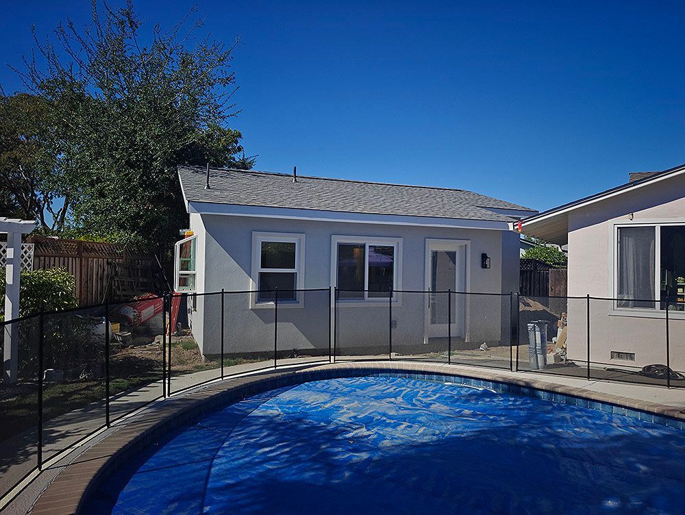 Backyard with a swimming pool in front of a gray building with windows and a door, sunny day.