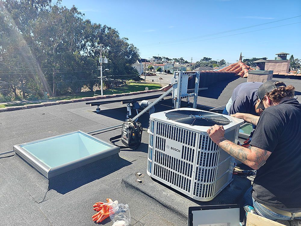 Two workers installing an air conditioning unit on a flat rooftop. Bright sunny day, cityscape in the background.