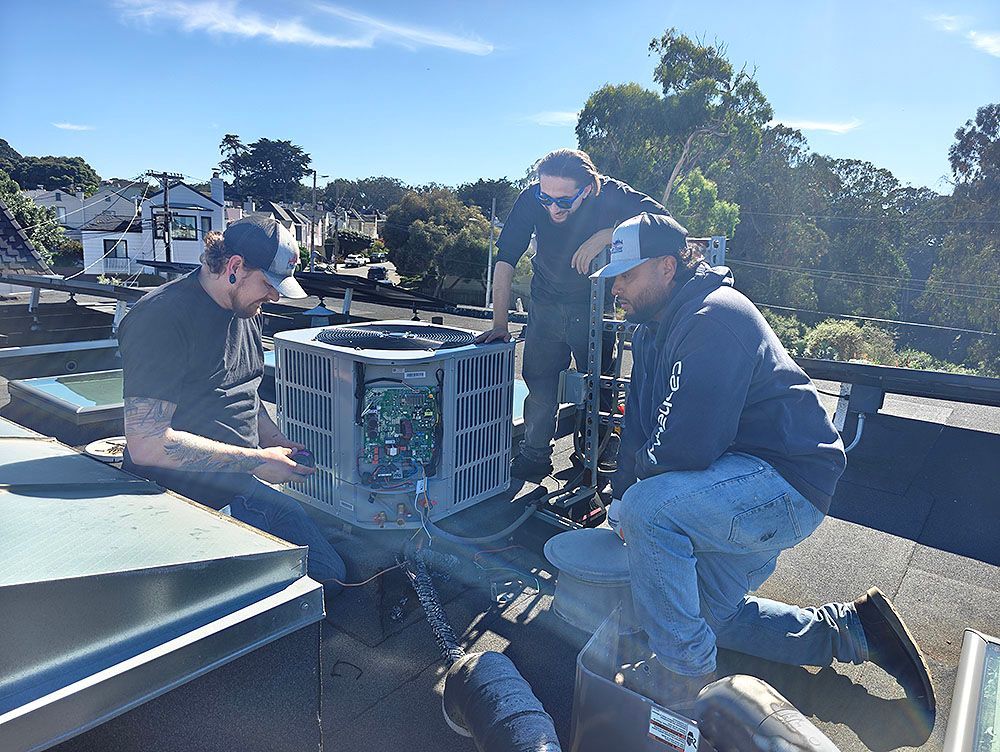 Three workers on a roof, inspecting an HVAC unit. Bright sunlight, blue sky, trees in the background.