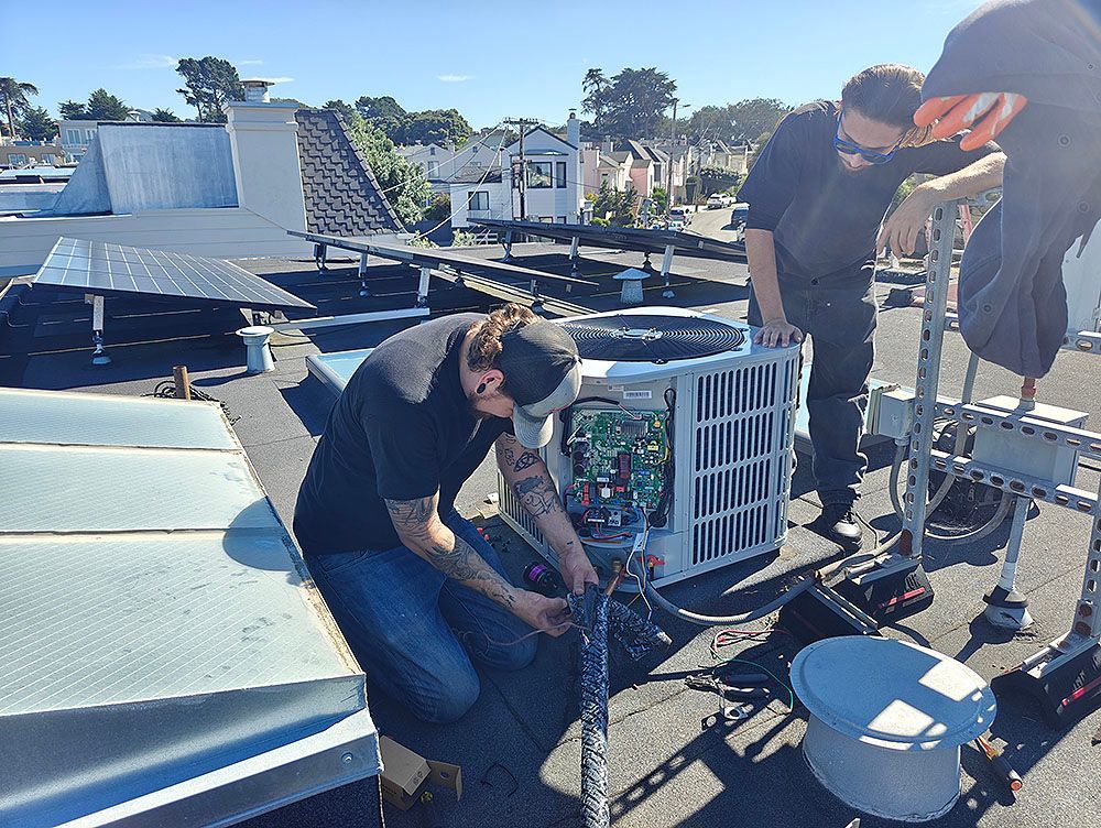 Three people working on an air conditioning unit on a rooftop. One man kneeling, working on wires.