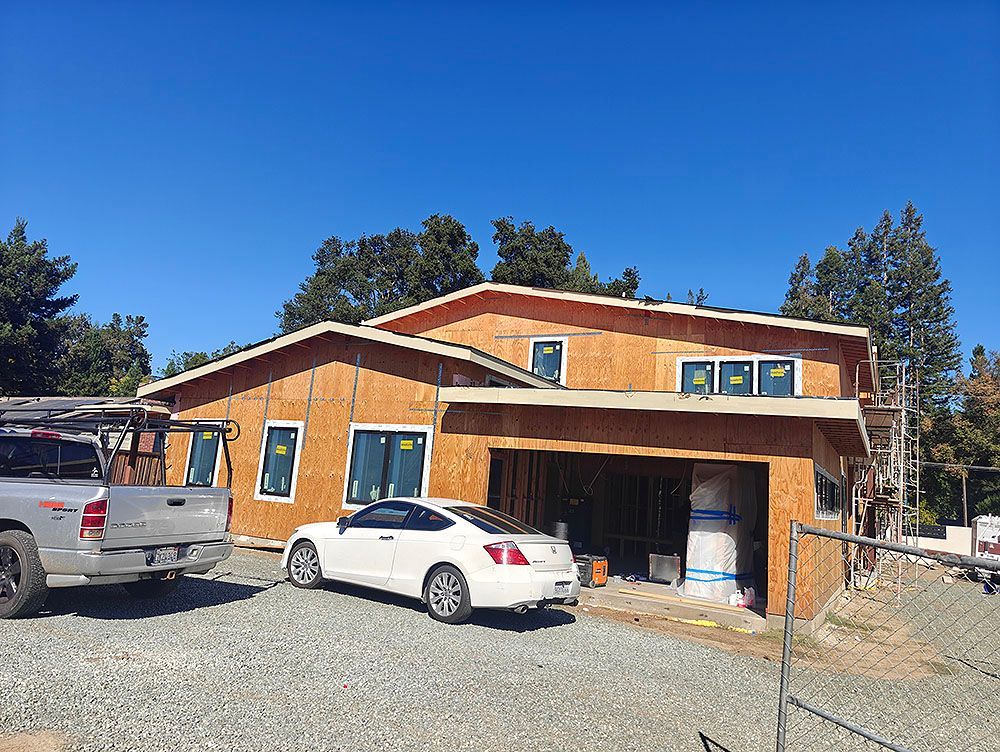 House under construction, with wooden frame, garage, and two parked cars on a gravel lot under a blue sky.