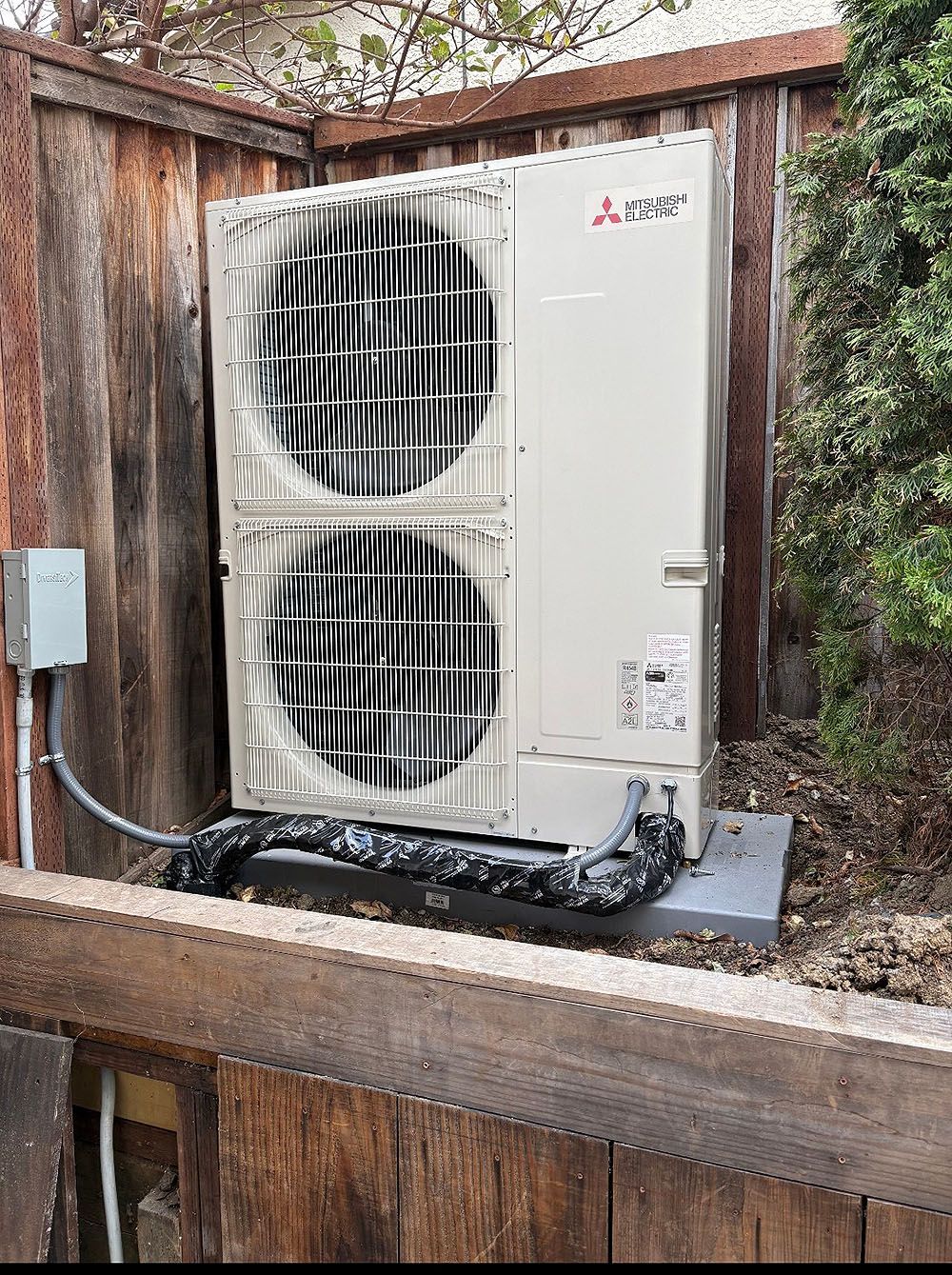 Mitsubishi heat pump against a wooden fence. Gray unit with two fans, black insulated pipes, and electrical box.