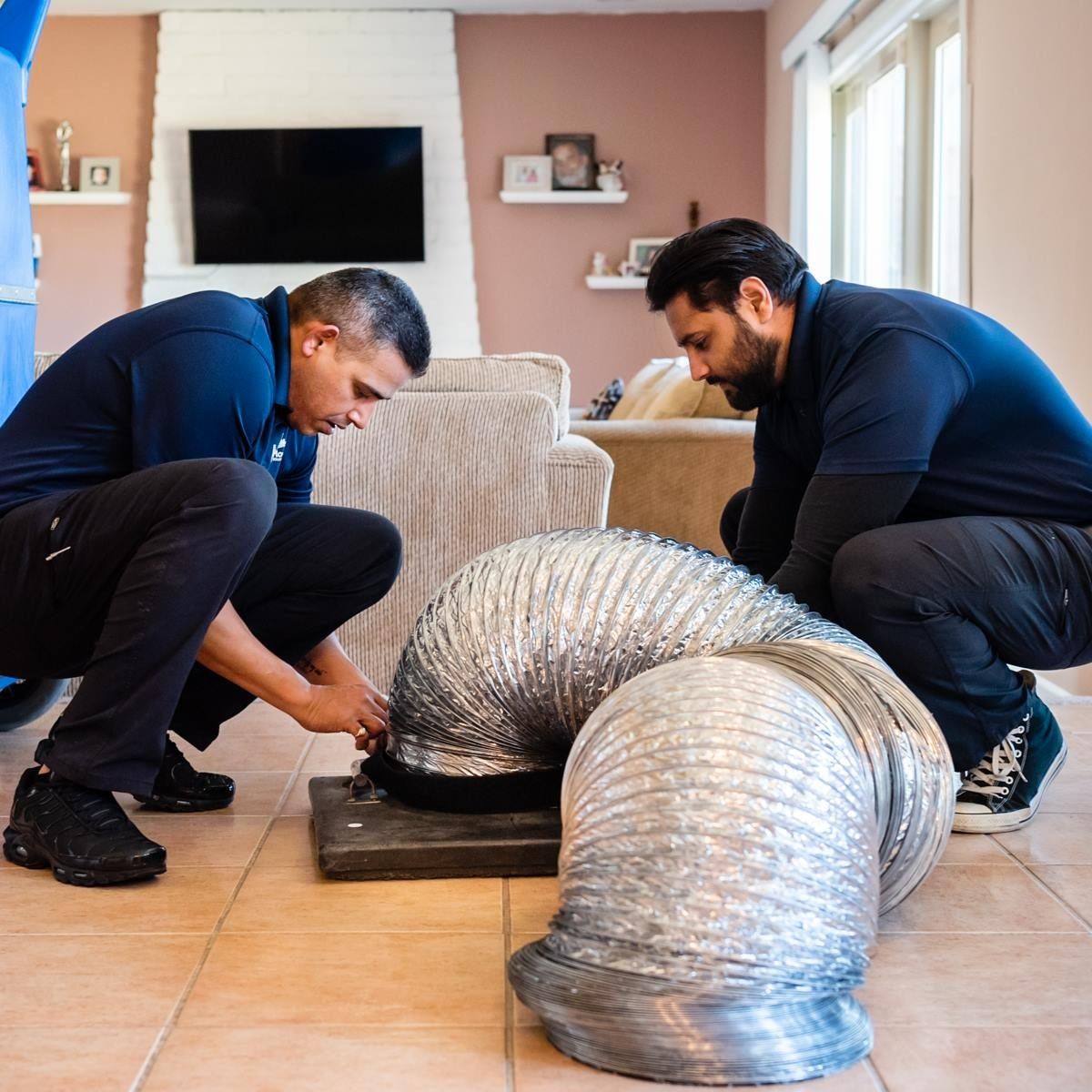 Two men in blue shirts, kneeling on the floor, working on a large silver ventilation duct in a home.