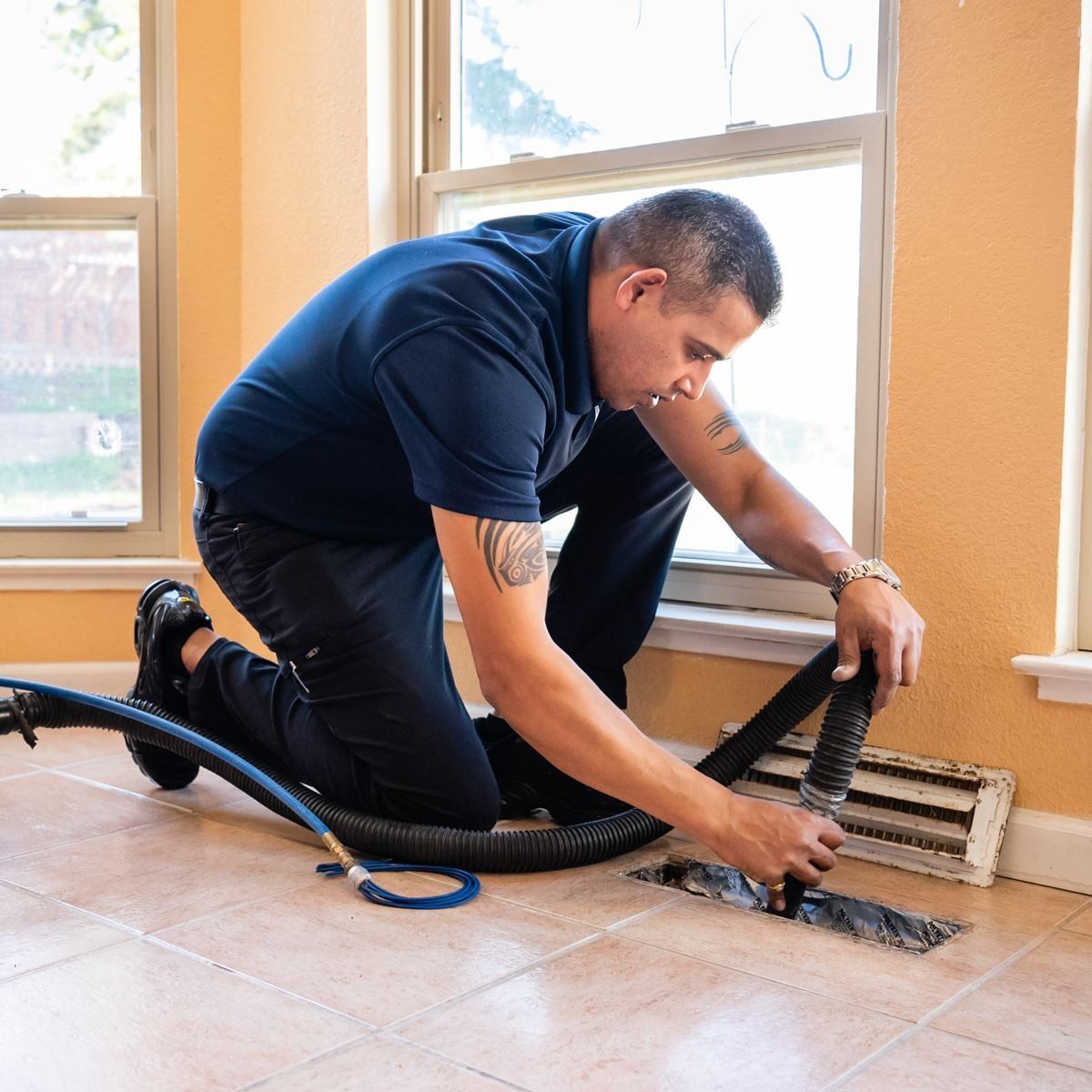 Man in blue shirt cleaning a floor vent with a hose in a room with windows.