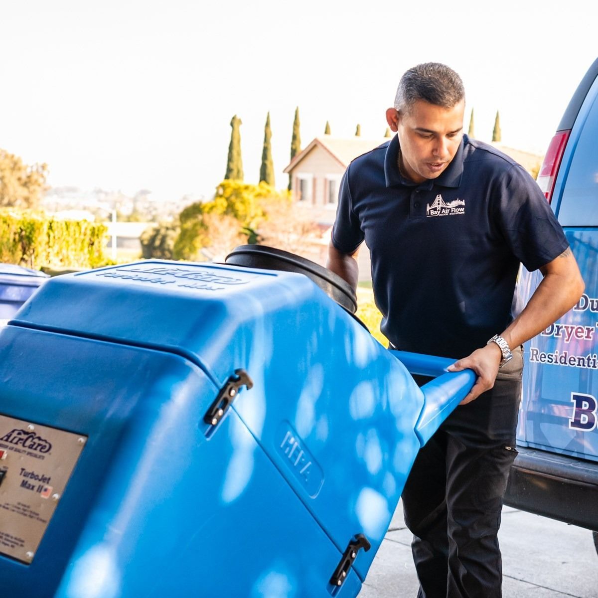 Man in uniform pulling a blue carpet cleaning machine from a van. Outdoors, daytime.