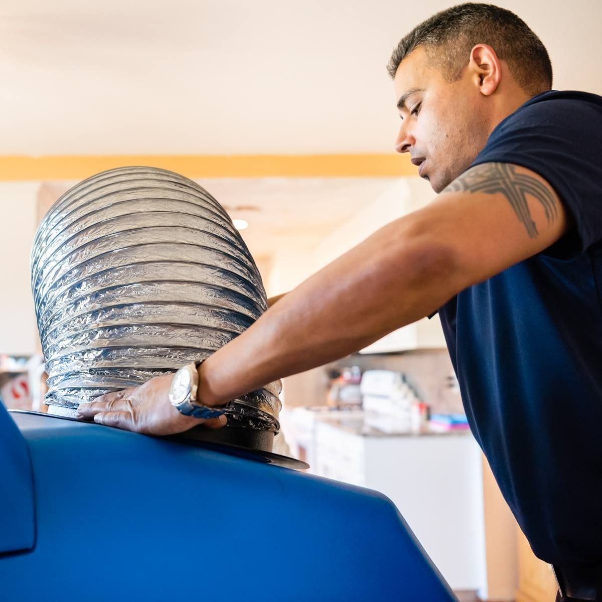 Man in blue shirt connecting flexible silver vent to blue machine indoors.