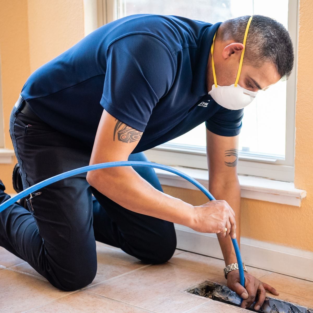 Man in mask cleaning vent in a home. He is kneeling and holding a blue hose.