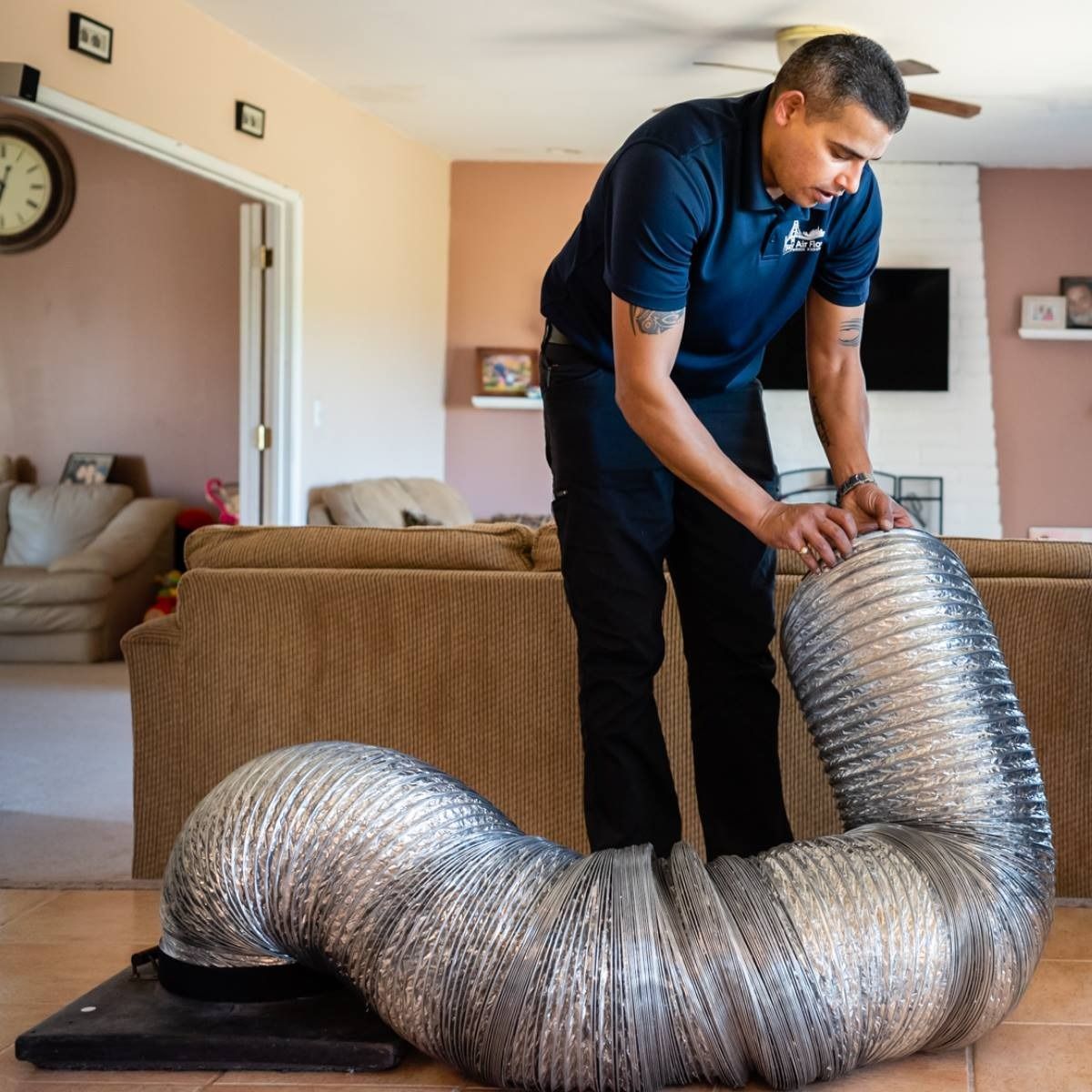 Man in a blue shirt inspects a silver flexible duct on a black base in a living room.