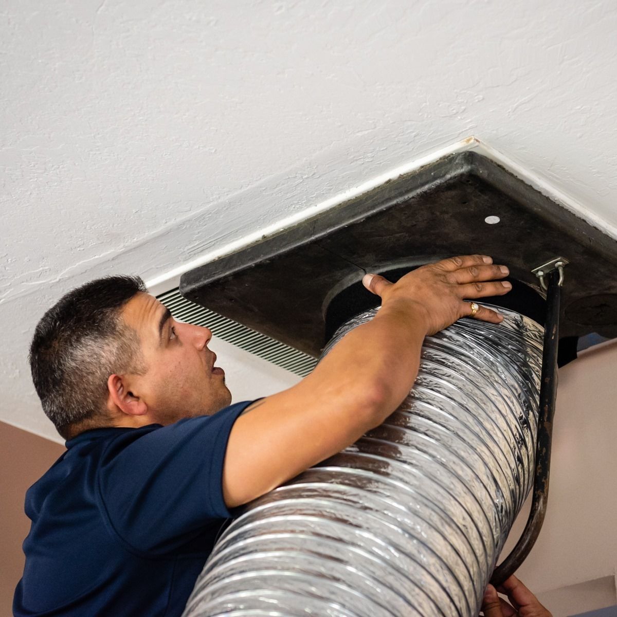 Man connects flexible silver duct to a vent in a white ceiling.