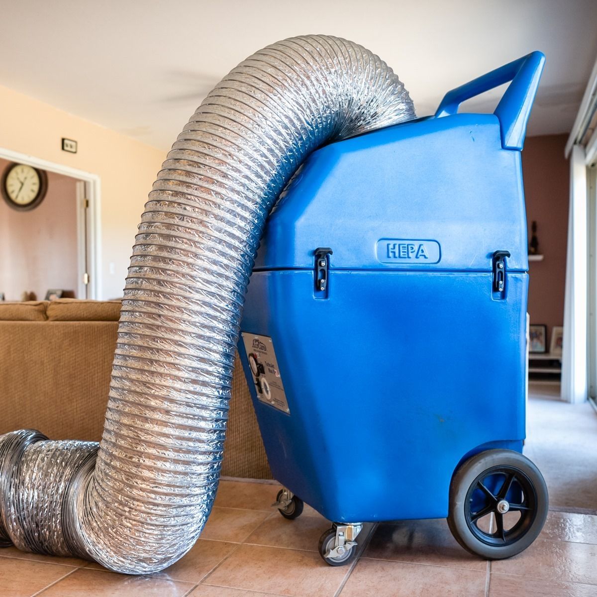 Blue HEPA air filtration machine with silver hose in a home.