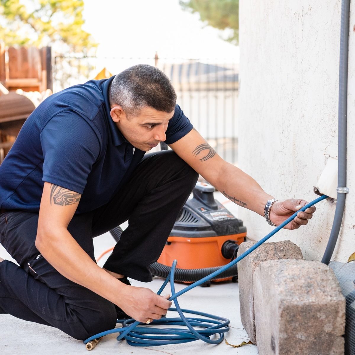 Man kneels to insert hose into wall outlet. Grey-haired, wearing dark shirt, pants. Vacuum cleaner nearby, outdoor setting.