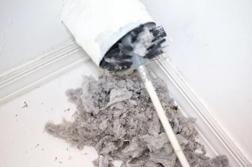 Brush cleaning a dryer vent, removing a pile of gray lint. White wall.