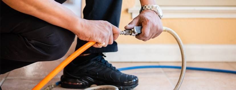 Person connecting an orange hose to a metal fitting, likely in a home. Wearing black pants and shoes, a watch.