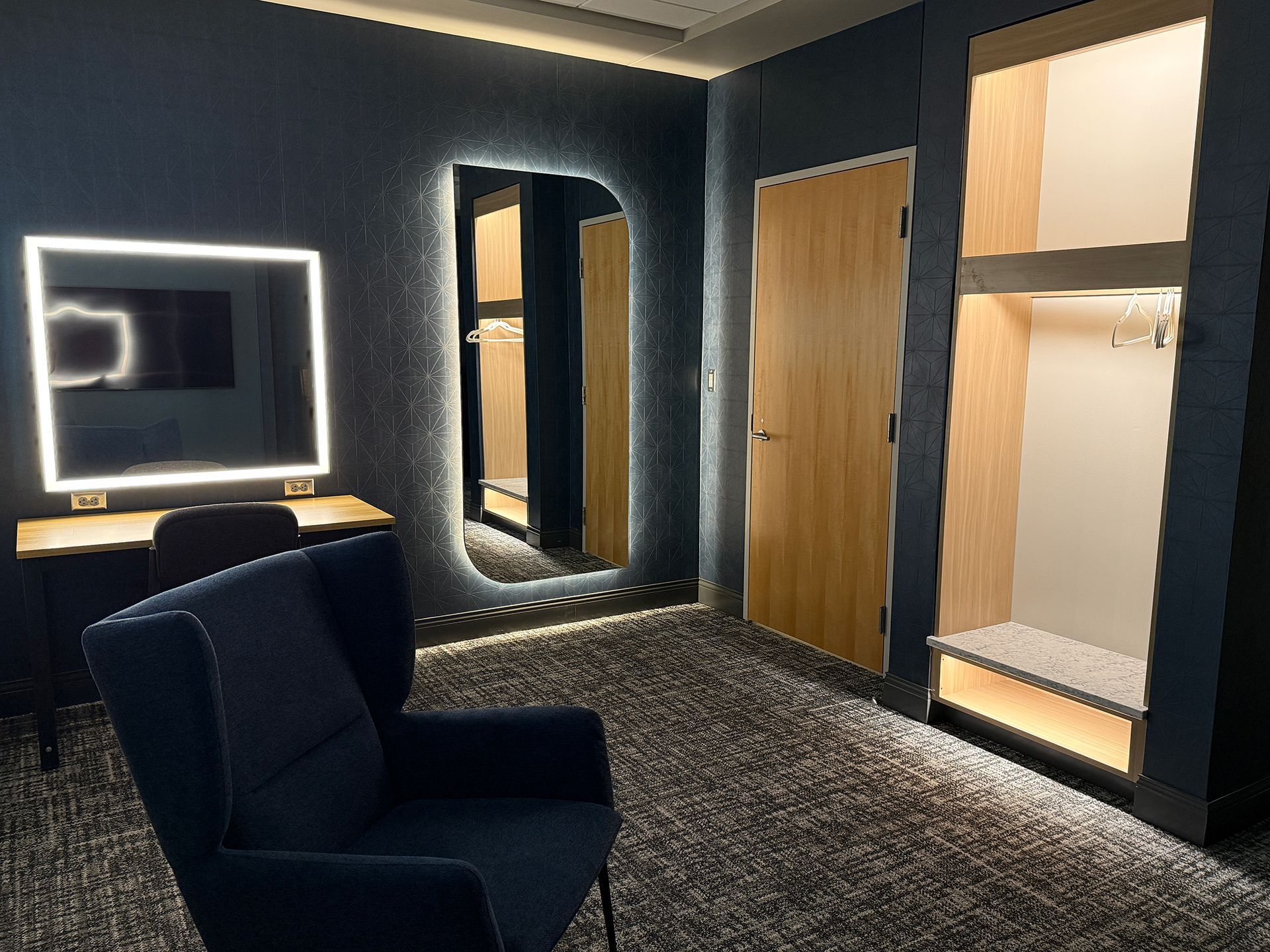 Modern hotel room with dark walls, plush chair, lit vanity mirror, and open closet with warm wood accents