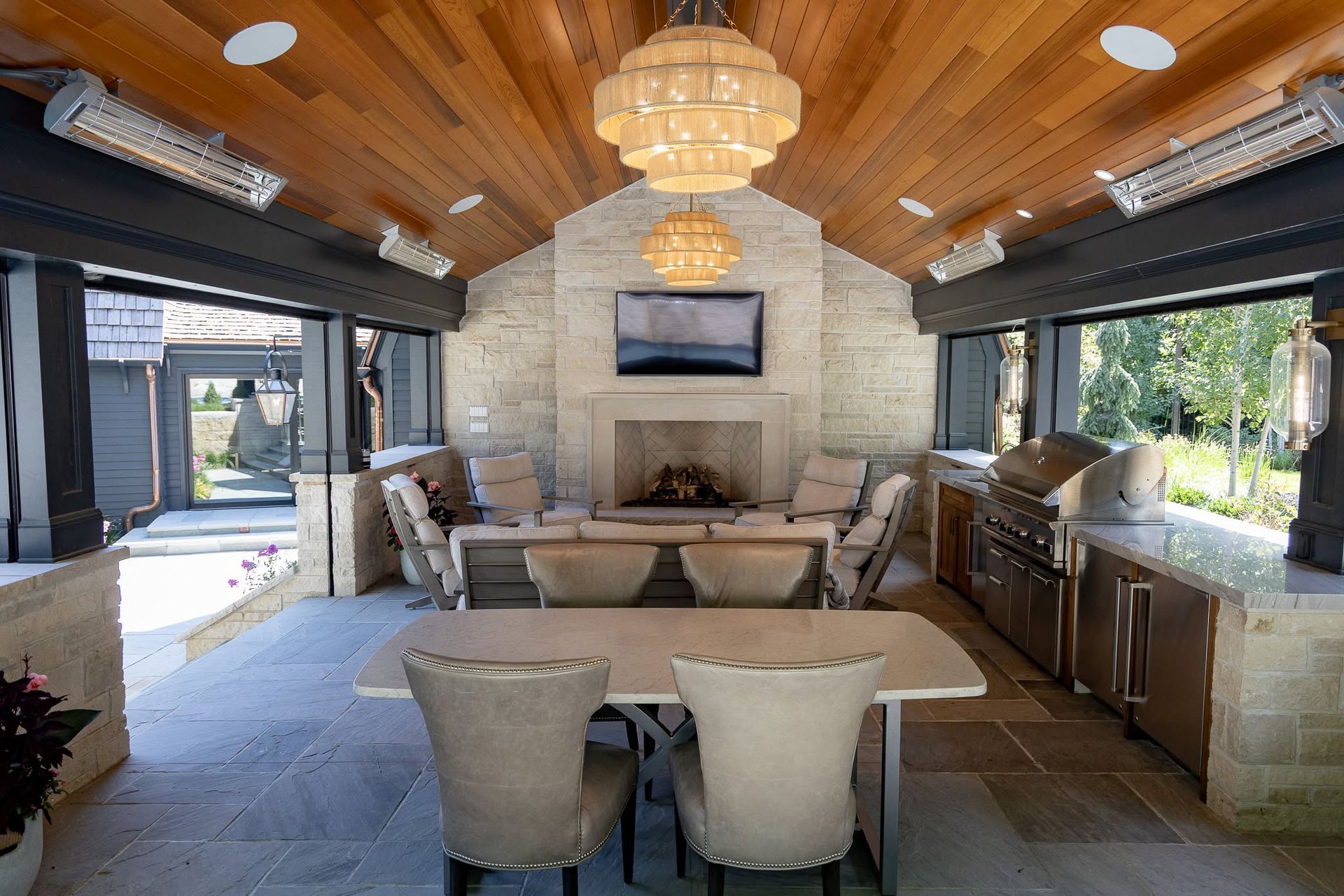 Open-air patio lounge with wood ceiling, fireplace, sofa, dining table, and large windows overlooking greenery