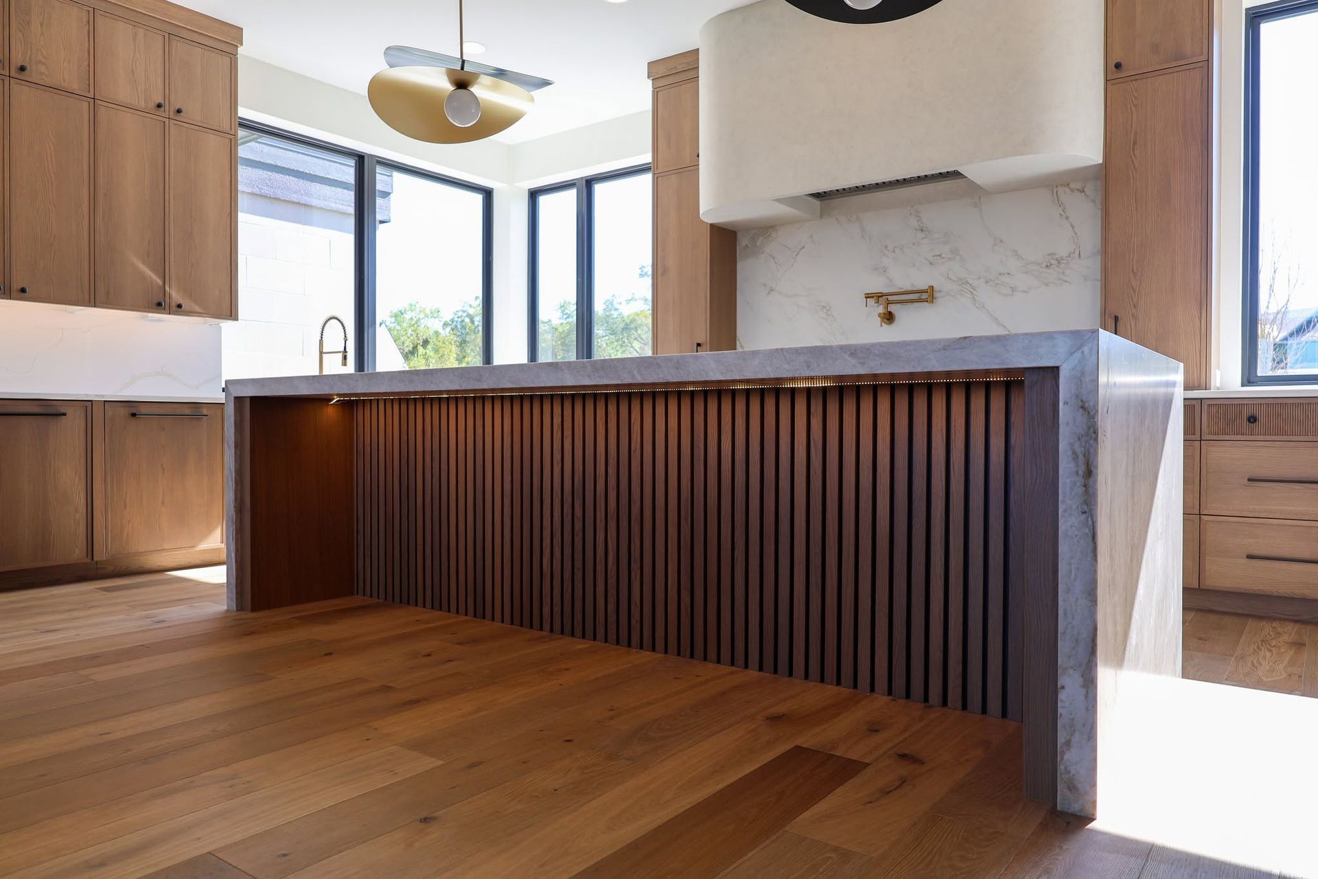 Modern kitchen with wood floors, dark slatted island, and large windows letting in daylight