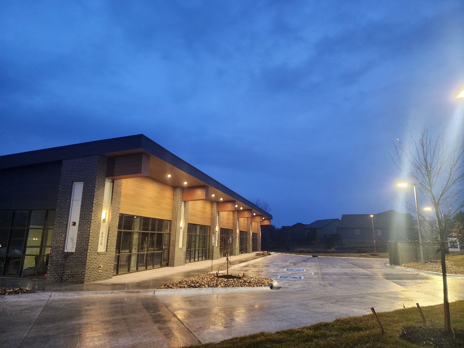 Modern building with warm lights at dusk, wet pavement reflecting the blue sky and streetlights.