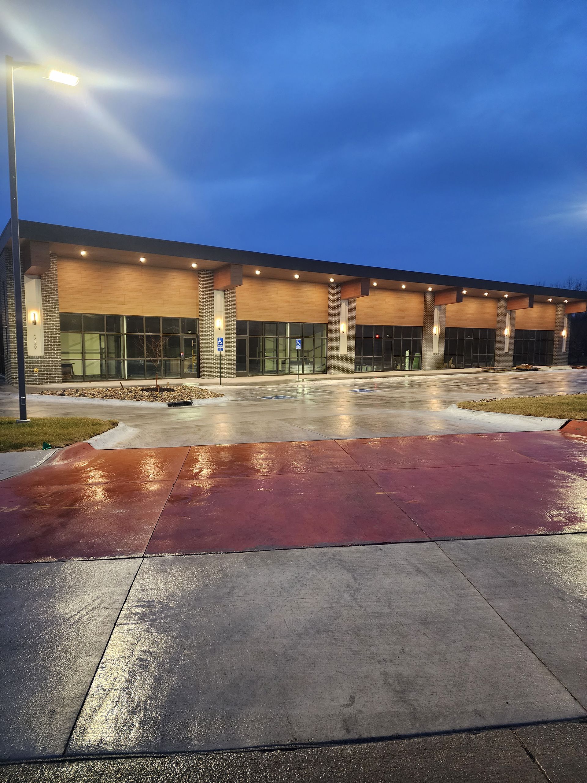 Rainy evening exterior of a modern building with lit windows, wet pavement, and a streetlight under a blue sky