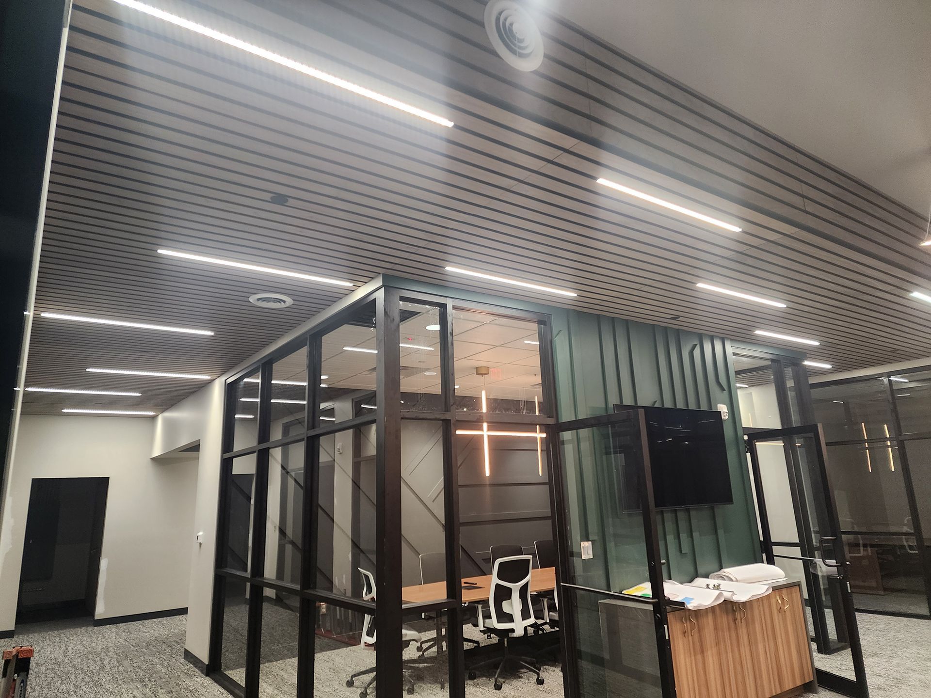 Glass-walled office interior with desks, monitors, and a striped ceiling under bright lights