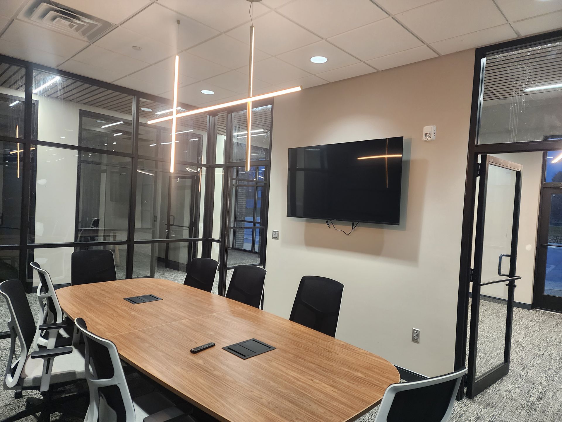 Modern conference room with a long wooden table, black chairs, glass walls, and a wall-mounted TV screen.