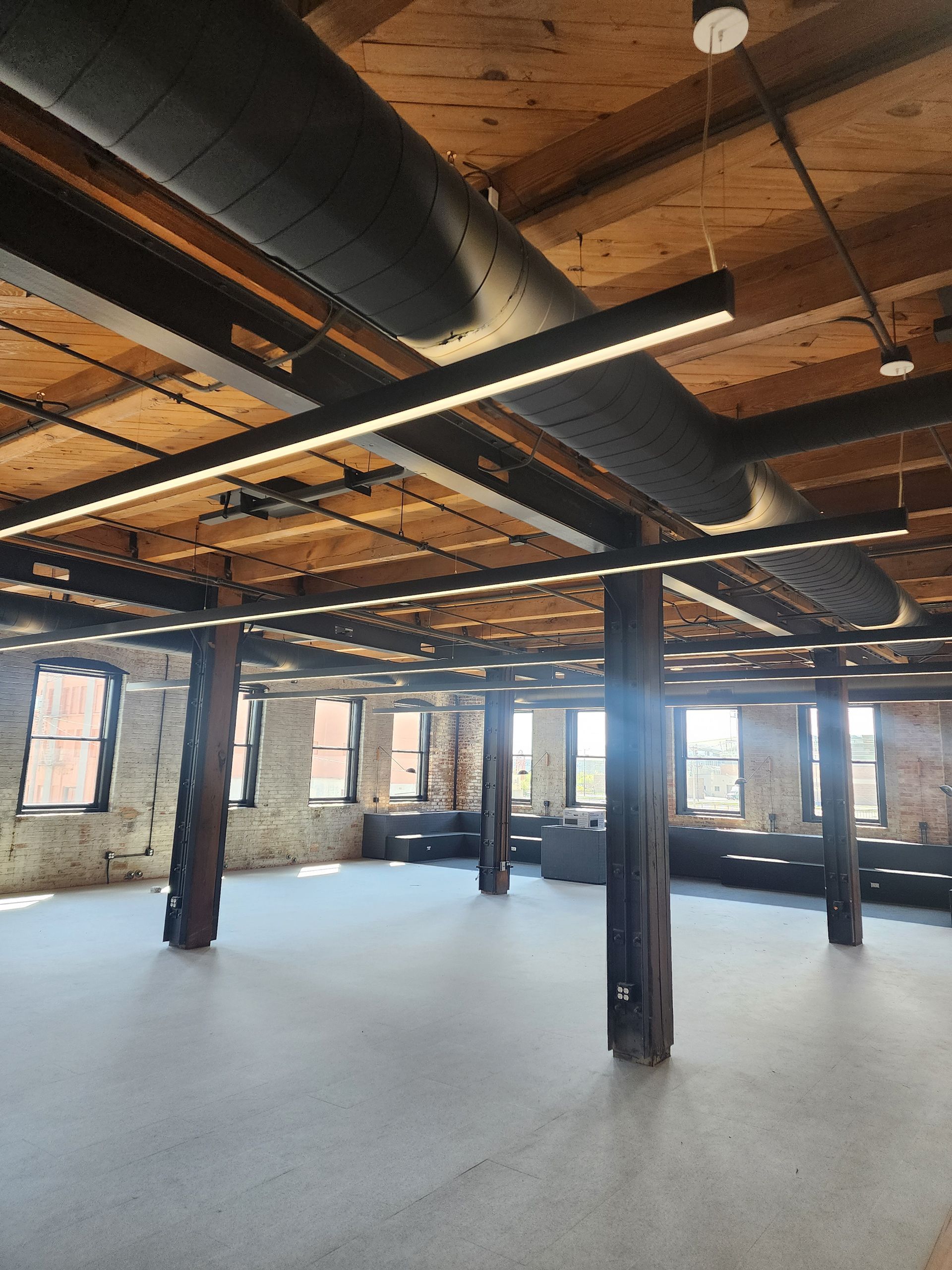 Empty loft interior with exposed brick columns, wooden ceiling beams, and large windows.