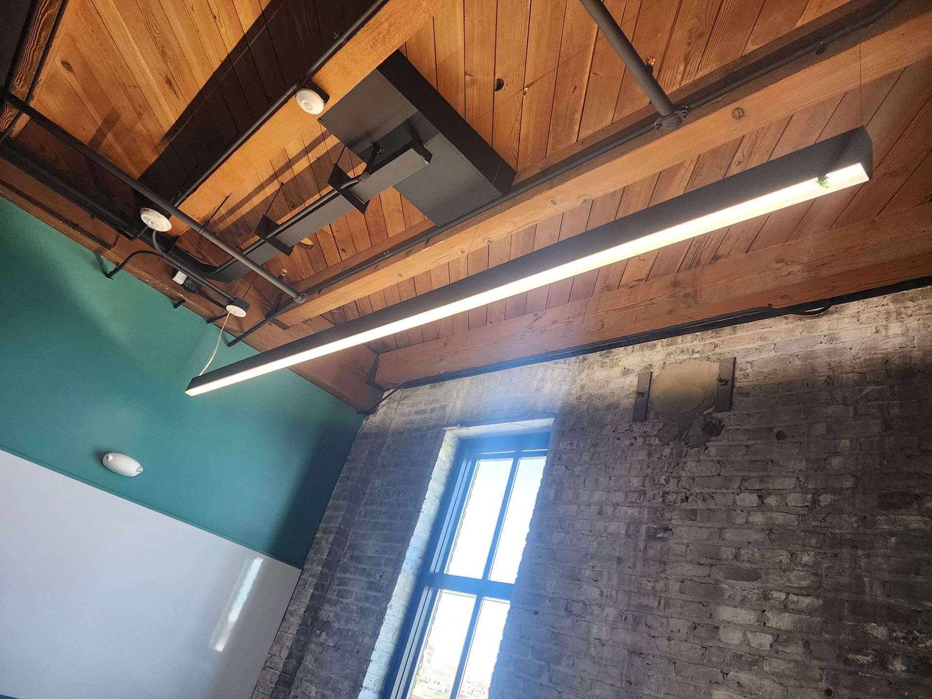 Industrial loft ceiling with exposed wood beams, hanging light fixture, and a tall window over a brick wall