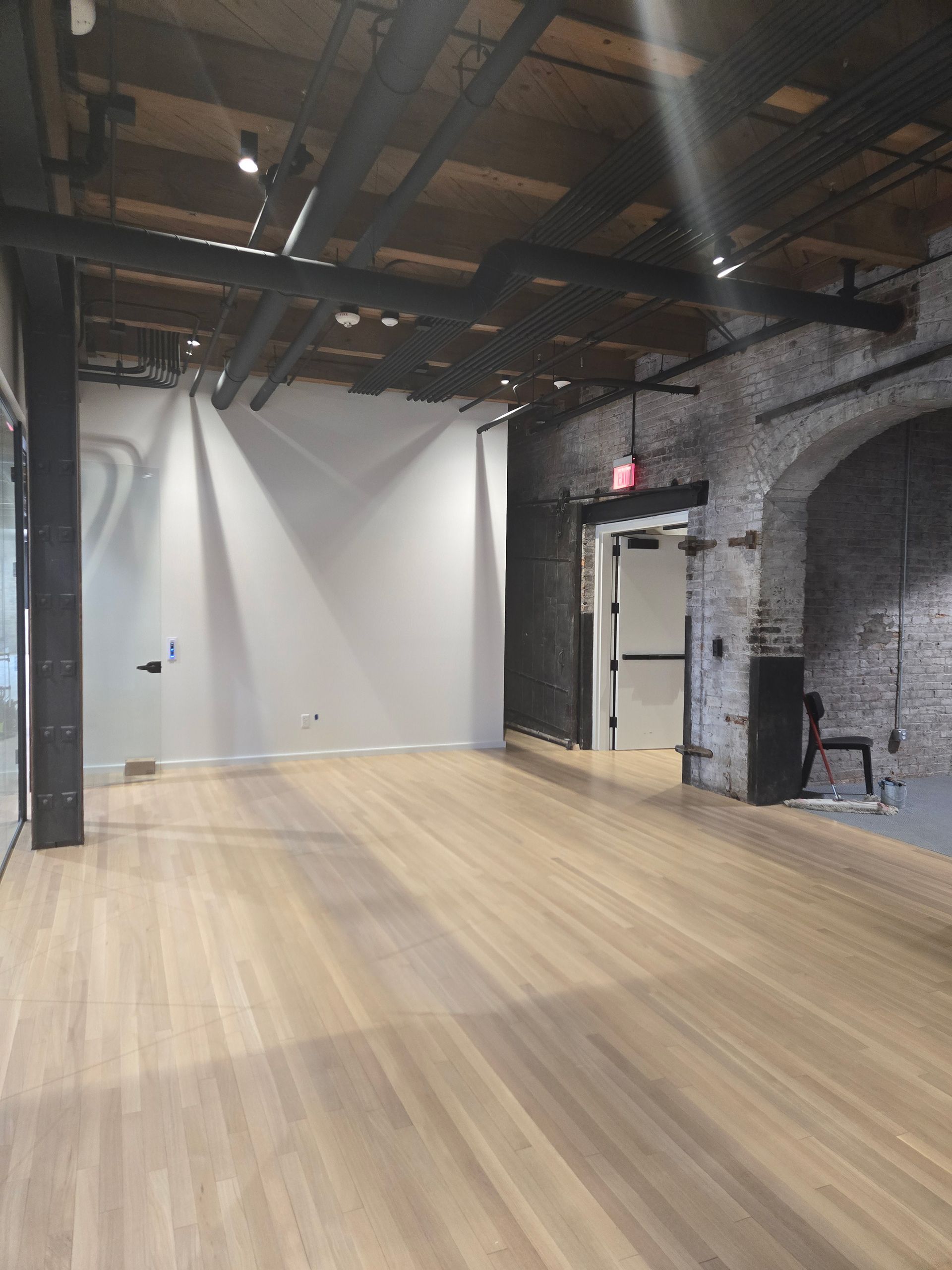 Empty industrial-style room with wood floors, white walls, exposed beams, and brick archway