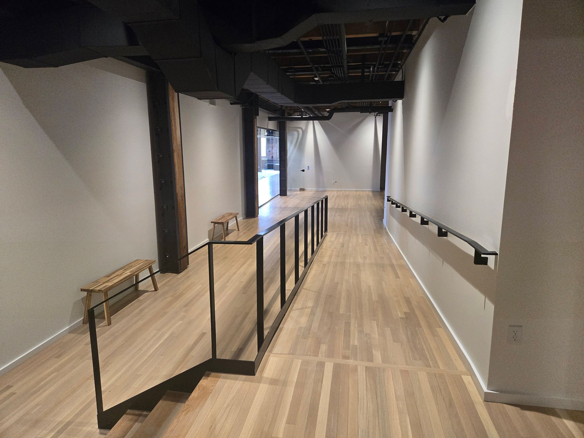 Empty indoor hallway with wood flooring, white walls, and black railings leading to a bright opening.